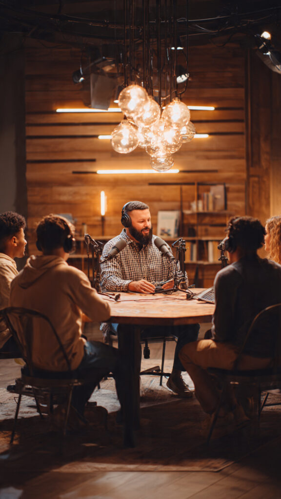 A pastor with beards, seated round a table, like podcast settings with some youths seated with him