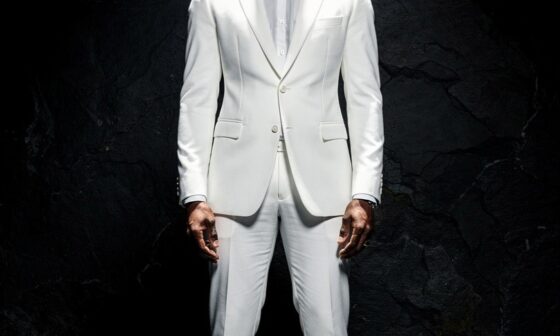 A full-body shot of a young black man standing upright in a brilliant, sparkling white suit, white socks, and white shoes inside a pitch-black environment under a single bright spotlight, symbolizing a firm stance of total structural immunity and purity.