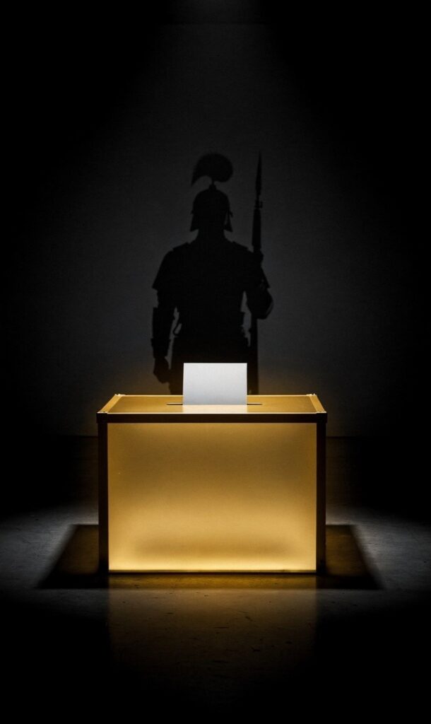 A dramatic, minimalist conceptual graphic showing a single voting ballot box glowing with crisp gold light, casting a long silhouette of a guard standing watch over a dark background.
